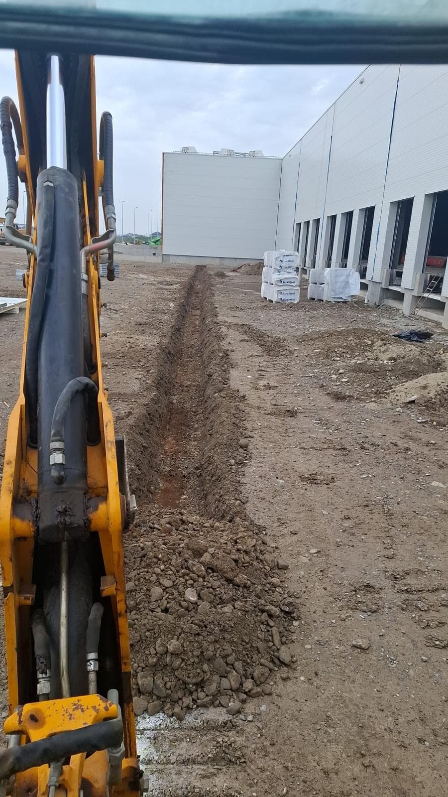 Excavator JCB săpând șanț de utilități la o hală industrială în Ilfov