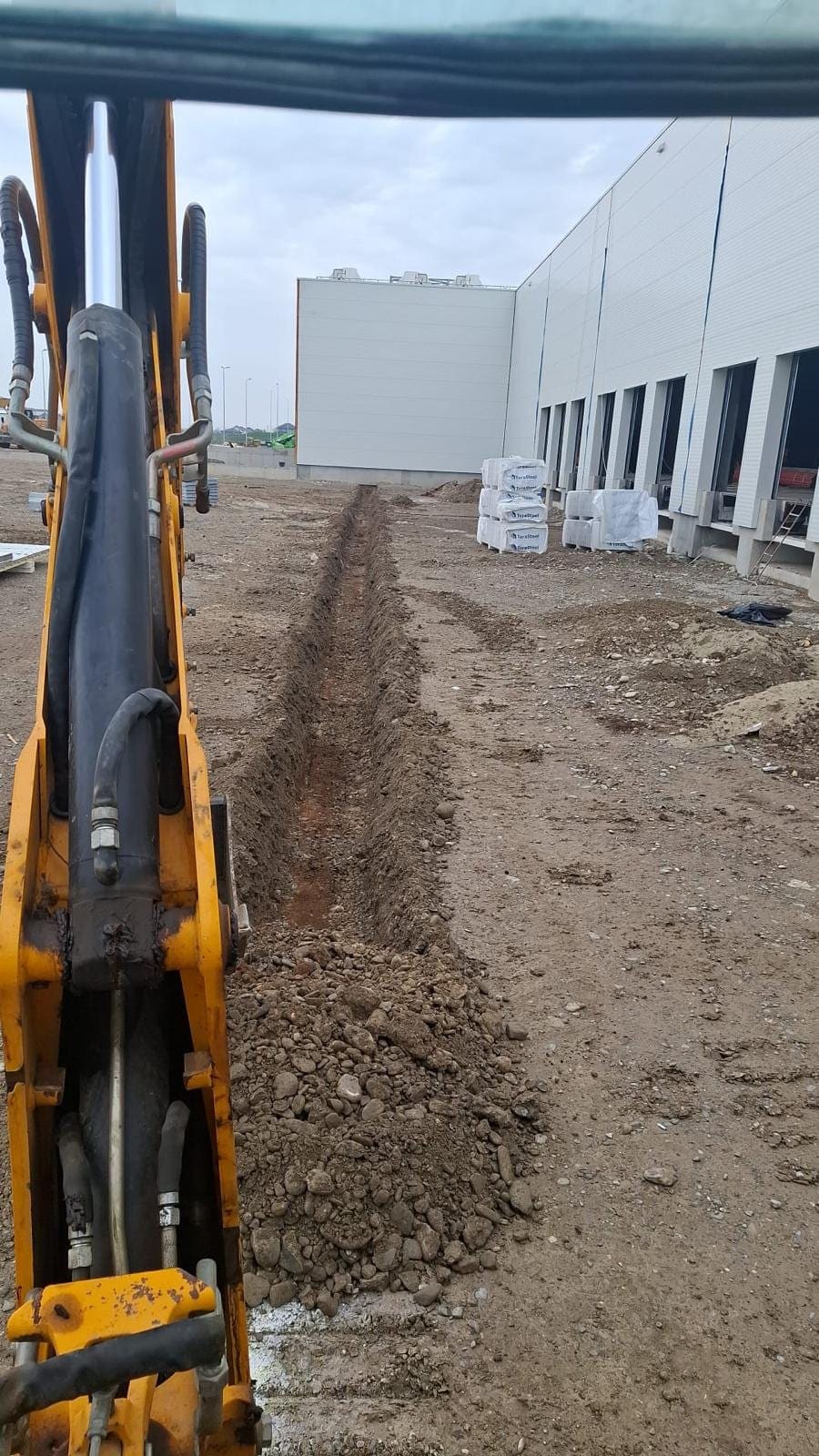 Excavator JCB săpând șanț de utilități la o hală industrială în Ilfov